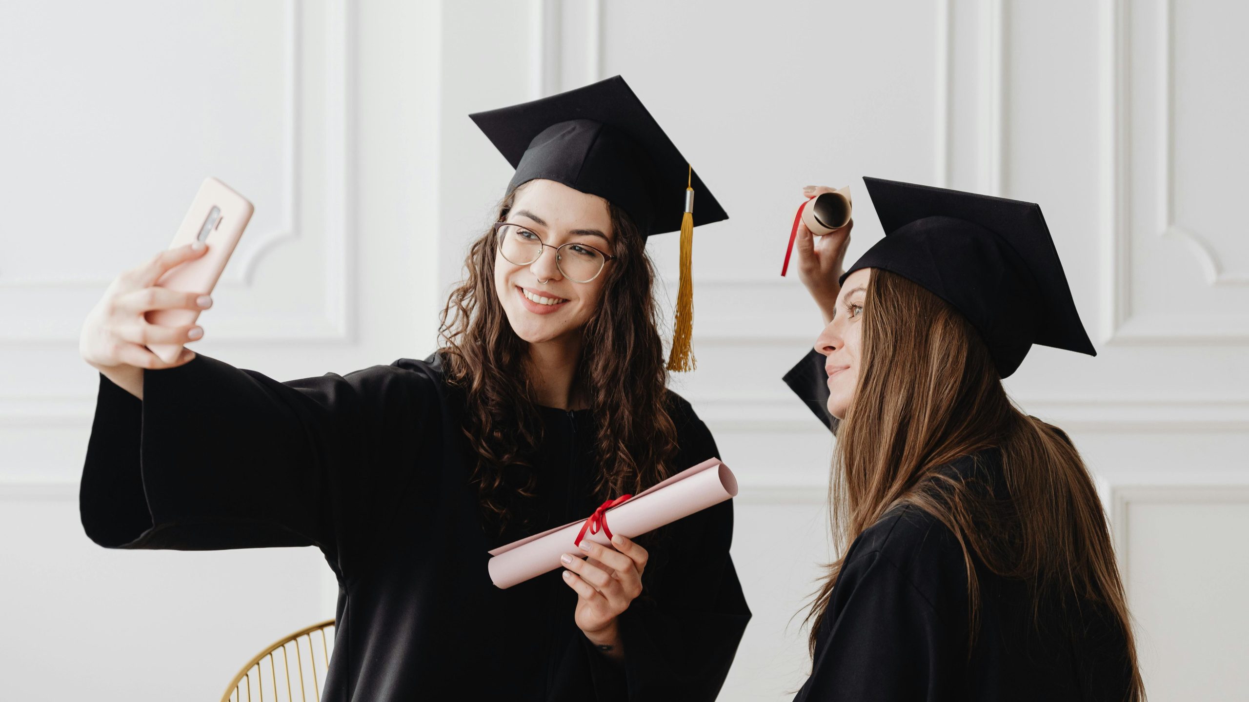 Two happy female graduates in academic gowns taking a selfie while holding diplomas indoors.